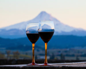 A pair of wine glasses in seen with red wine in the foreground with Mt Hood at sunset with an alpine glow in the background near Hood River, OR.